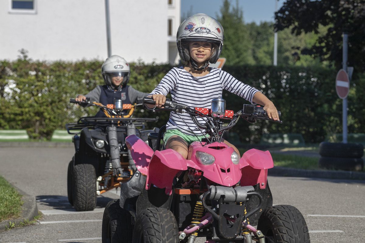 Mädchen mit Helm auf rosa Kinderquad mit Jungen auf Quad im Hintergund